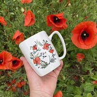 biały kubek z namalowanymi w kółku makami i napisem I'love You, na tle kwitnących maków